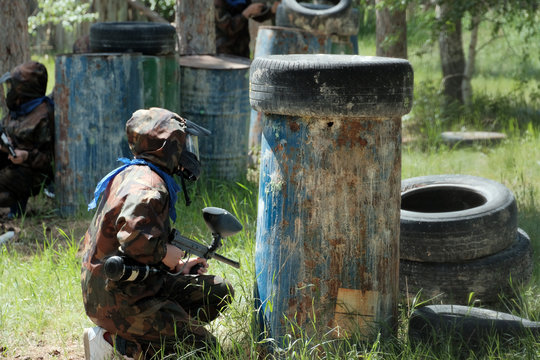 Paintball. Players in camouflage clothing with weapons in their hands. Waiting in ambush in the forest among trees, iron barrels, car tires. Team play.