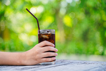 cola and ice in glass with straw in hand on blurred tree background