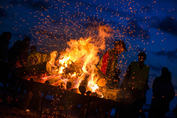 Large burning bonfire with soft glowing flame and sparkles flying all around. Romantic summer evening, people relaxing and enjoying calmness at the seaside during the Night of ancient lights.   - obrazy, fototapety, plakaty