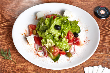 salad with mozzarella cheese from cucumbers, tomatoes, olives, bell peppers, onion rings, lettuce, lies on a white plate decorated with red pepper