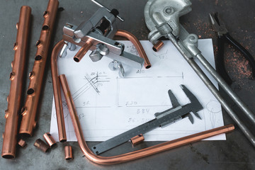 Brass pipes, pipe bender, pipe rammer tool and a layout drafts on a fitter workbench.