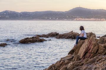 Fototapeta na wymiar Young woman sitting on the rock by the seaside and dreamily watching the sea and coastline after sunset.