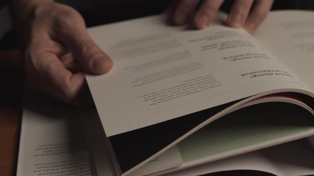 Person flipping through book seeking inspiration on a wood table in a design studio. Dolly shot, moody lighting.