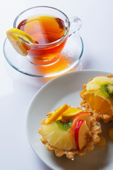 Basket-cake decorated with different fruit and cup of tea on white table.