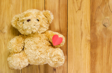 Teddy bear holding red paper heart shape on wooden background.