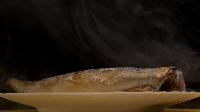 Plate with frozen mackerel fish on a black background from which there is frosty freshness and evaporation, close-up, 4K