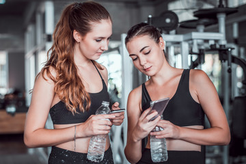 Two young attractive woman talking together and using smart phone or cellphone while relaxation after hard workout at gym