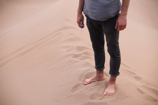 Man with earphones in his hand is standing barefoot on a top of a sand hill in big beautiful orange desert sand dunes in Yazd, Iran