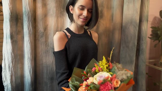 Sexy brunette in black shoulderless sweater standing with big bouquet of flowers. Girl posing for camera happy, satisfied and smiling, adoring flowers, brushing her hair behind ear, looking at flowers