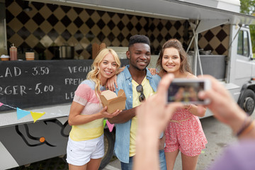 leisure, technology and people concept - young man taking picture of his happy friends eating hamburgers and wok at food truck