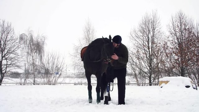 winter, disabled man stands near black horse on snowy field. man strokes a muzzle of a horse. man has prosthesis instead of his right leg. he learns to ride horse, hippotherapy. rehabilitation of