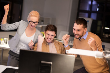 business, success and teamwork concept - happy coworkers with computer celebrating victory at night office