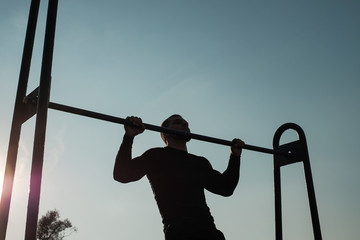 A young muscular man pulls himself up on a bar in the background of the sun on the street.