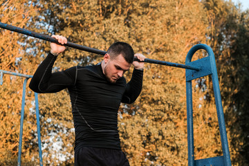 A young muscular man pulls himself up on a bar in the background of the sun on the street.