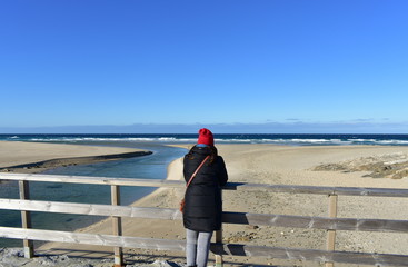Beach with woman resting on a wooden handrail and looking at the view. Winter clothes and red hat....