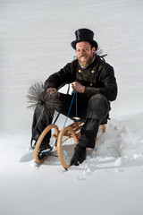 Schornsteinfeger in Arbeitskleidung, mit Kehrgeräten , sitzt auf Holzschlitten im Schnee