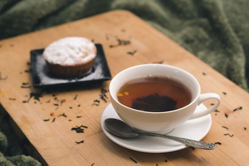 Homemade cupcake with powdered sugar on a black plate and a white cup of tea with natural additives on a wooden tray on a background of green textiles. The concept of a cozy breakfast in bed