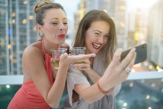 Two Beautiful Friends Taking Selfie On Rooftop Bar With City Lights Backdrop