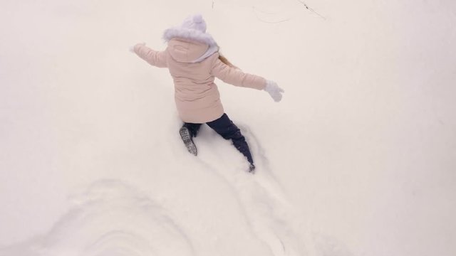 Young girl in coral pink jacket walk and fall to snow than show snow angel butterfly movement of legs and hands overhead view top to down snowfall play. Slow motion. Girl lies at white snow