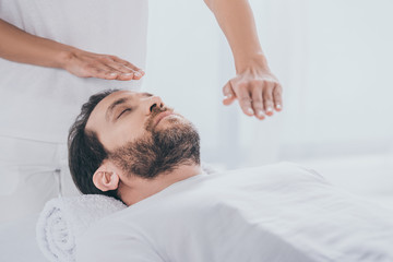 calm bearded man lying on massage table and hands of healer doing reiki treatment session