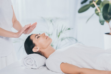cropped shot of peaceful young woman with closed eyes receiving reiki treatment on head