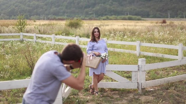 Photographer make a photo for beautiful pregnant woman dressed in the blue dress and knitted hat. She put hands on her belly. Photosession in the field near the farm. Summer time