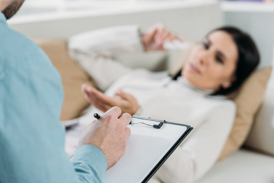 cropped shot of psychotherapist writing on clipboard and young patient in depression lying on couch