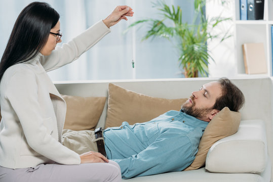 side view of young hypnotist with pendulum and bearded man with closed eyes lying in couch during hypnotherapy