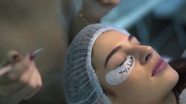Woman is in cosmetologic salon.She is lying with a closed eyes and in hair mask. A beautician is doing the procedure of eyelash extension.