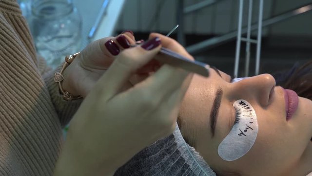 Woman is in cosmetologic salon.She is lying with a closed eyes and in hair mask. A beautician is doing the procedure of eyelash extension. With special equipment she is styling lashes.