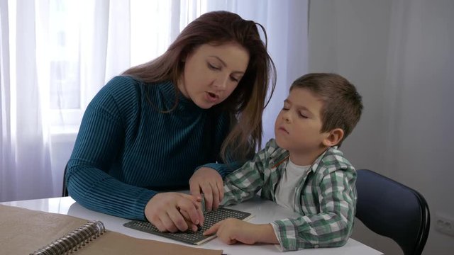 education Visually impaired children, mom teaches blind son boy to write braille characters font sitting at table in bright room at home