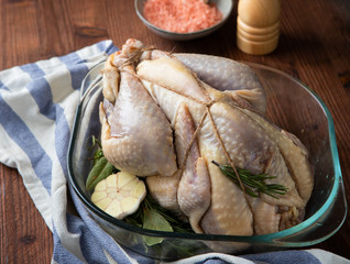 Raw guinea fowl in transparent glass baking dish with garlic and rosemary and next to it are Himalayan pink salt in a bowl and a pepper mill