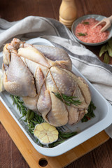 Raw guinea fowl in a white baking dish with garlic and rosemary and next to it are Himalayan pink salt in a bowl and a pepper mill