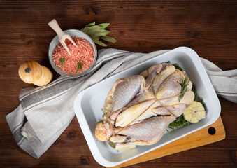 Raw guinea fowl in a white baking dish with garlic and rosemary and next to it are Himalayan pink salt in a bowl and a pepper mill