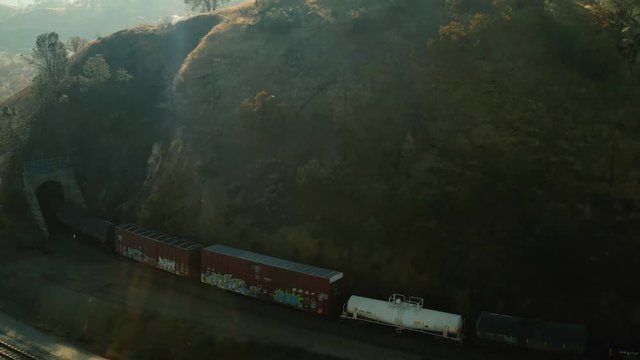 The aerial camera come flying in at 60 feet flying sideways, panning to the right and accelerating past the rail cars then to follow the three locomotives into the southern entrance of the rail loop.