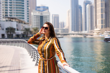 Happy beautiful tourist woman in fashionable summer dress and sunglasses walking in Dubai marina in United Arab Emirates. Luxury and comfortable tourism season in UAE. Warm summer day.