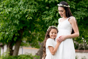 happy pregnant mother and cute smiling daughter in white dresses and wreaths standing and hugging in park