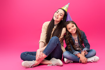 full length view of happy mother and daughter in party hats sitting together and smiling at camera on pink