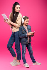 Fototapeta premium full length view of smiling mother holding digital tablet and looking at daughter in headphones using smartphone on pink