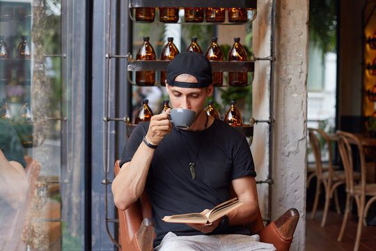Relaxed young bearded Caucasian guy enjoys spare time, calm atmosphere outdoors, holds mug with coffee, reads interesting novel, looks attentively at book, sits armchair over stylish cafe interior.