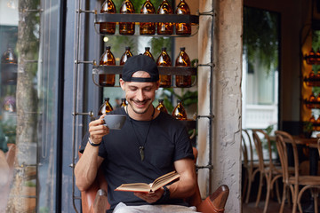 Handsome stylish guy holds interesting book, sits in modern restaurant, wears casual stylish clothes, laughs out load over cafe background. Positive man reads pop literature with good sense of humor.