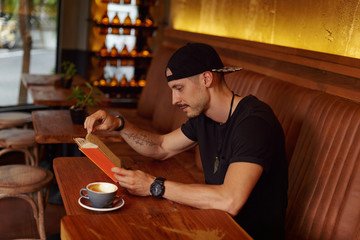 Bearded young guy reads scientific literature, enjoys moment to be alone in cafe with fresh coffee, reads foreign book, sitting in modern cafe with stylish interior at wooden table. Education concept.
