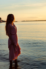 Beautiful woman walking on water at sunset on sea beach.  Young woman wearing long beige dress standing on  beach at sea.