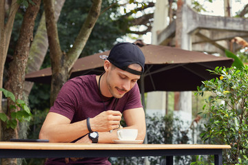Young male hipster having coffee during lunch break while sitting at wooden table at restaurant. Handsome trendy guy holding spoon to taste coffee foam, enjoying good morning, breath fresh air outdoor