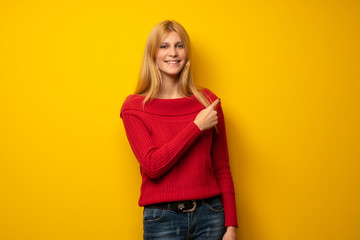 Blonde woman over yellow wall pointing to the side to present a product