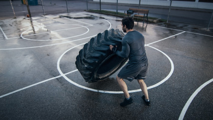 Strong Muscular Fit Young Man in Sport Outfit and Gloves is Doing Exercises in a Fenced Outdoor Basketball Court. He's Flipping a Big Heavy Tire in an Afternoon Environment After Rain. Backshot View