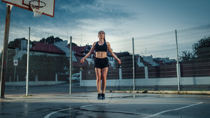 Beautiful Energetic Fitness Girl Skipping/Jumping Rope. She is Doing a Workout in a Fenced Outdoor Basketball Court. Evening Shot After Rain in a Residential Neighborhood Area. - obrazy, fototapety, plakaty