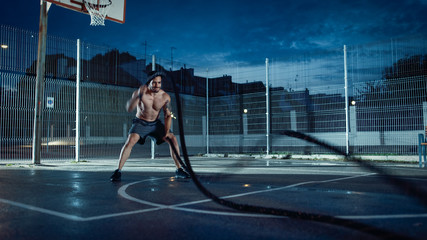 Strong Muscular Fit Shirtless Young Man is Doing Exercises with Battle Ropes. He is Doing a Workout in a Fenced Outdoor Basketball Court. Evening After Rain in a Residential Neighborhood Area.