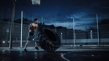 Strong Muscular Fit Young Shirtless Man is Doing Exercises in a Fenced Outdoor Basketball Court. He's Flipping a Big Heavy Tire at Night After Rain in a Residential Neighborhood Area.