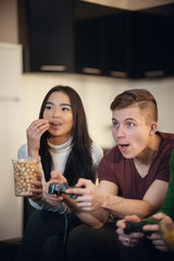 Company of young friends spending time together playing games and eating food. Young man playing game and his girlfriend watching